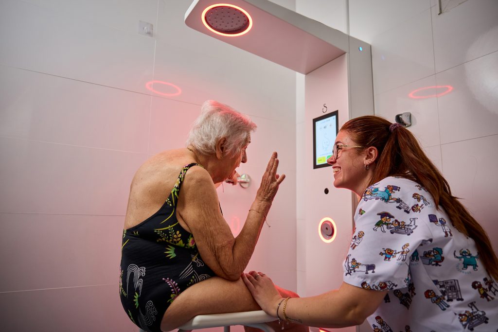 Older woman assisted by a care professional while using the Showee smart shower in an accessible bathroom.